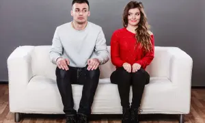 Coppia seduta su un divano bianco, lui guarda altrove con espressione incerta, lei sorride. Come superare la timidezza in amore?