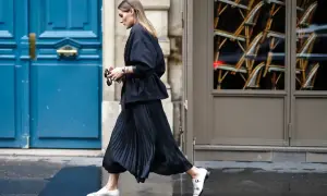 Elegancka kobieta w czarnej, plisowanej spódnicy i marynarce, tworząca stylową stylizację. Białe mokasyny dodają kontrastu.