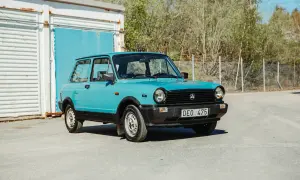 En ljusblå Lancia A112 Junior står parkerad framför en blå port och en vit containerbyggnad.