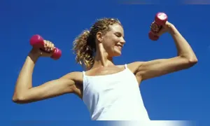 Femme souriante avec des haltères roses, montrant ses muscle bras. Elle porte un débardeur blanc sous un ciel bleu clair.