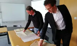 Deux hommes consultent des registres d'état civil aux archives départementales du Morbihan. L'un pointe un document ancien.