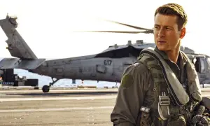 Glen Powell w filmach jako pilot na pokładzie lotniskowca, z helikopterem w tle.