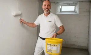 Mann streicht Kellerwand im Altbau mit weißer Farbe. Er hält eine gelbe Farbeimer-Marke "StoColor Sil In".