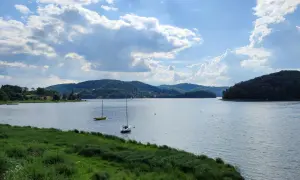 Widok na jezioro z żaglówkami i zalesionymi wzgórzami w tle, przy słonecznej pogodzie z chmurami.