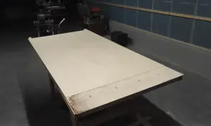 Stor skiva av plywood på ett arbetsbord, redo att bygga slagport. Verktyg syns i bakgrunden.