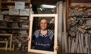 Mujer sonriente en taller, rodeada de rollos de tela y estanterías con materiales. Sostiene un marco de madera en rollo.