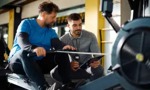 Dwóch mężczyzn na siłowni, jeden ćwiczy na wioślarzu, drugi z notatnikiem. Rozmawiają o tym, ile zarabia instruktor fitness.