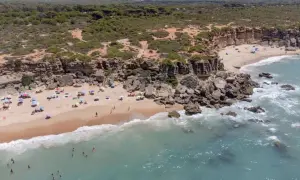 Una cala enebro con acantilados rocosos, arena dorada y aguas turquesas. Gente disfrutando del sol y el mar bajo sombrillas.