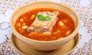 Aromaticzna zupa na żeberkach z fasolą i ziołami, podana w glinianej misce na ozdobnym obrusie.