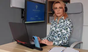 Kobieta w okularach i słuchawkach siedzi przy biurku, pracuje na laptopie. Na monitorze widnieje logo i dane kontaktowe.