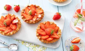 Trois tartelettes aux fraises, garnies de crème et de fruits rouges frais, sur un plateau blanc. Une fraise entière repose à côté.