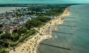 Tłum ludzi na plaży w Mielnie, cieszących się słońcem i morzem. W tle widać zabudowania i jezioro. To idealne miejsce na wakacje, pełne atrakcji.