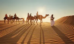 Kamelkarawane in der Wüste bei Sonnenuntergang. Ein Führer in traditioneller Kleidung begleitet Touristen. Tolle Abu Dhabi Tipps für ein unvergessliches Erlebnis.