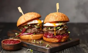 Dwa soczyste hamburgery z grilla, z jajkiem sadzonym i sosem, prezentują się apetycznie na drewnianej desce.