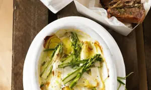 Überbackener Spargel mit Kartoffeln und Parmesan in einer Auflaufform, daneben ein Stück Lammkotelett.
