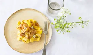 Ein Teller mit cremiger Pasta, die wie Fettuccine aussieht, garniert mit zarten Blüten und Speck. Daneben ein Glas Wasser und ein Strauß weißer Blumen.