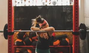 Erschöpfte Athletin lehnt an Hantelstange. Eine Woche Trainingspause ist sinnvoll für Regeneration und neue Motivation.