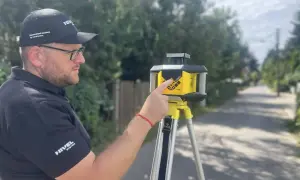 Mężczyzna w czapce z daszkiem i okularach obsługuje niwelator laserowy na statywie. To urządzenie pomaga w precyzyjnych pomiarach budowlanych.