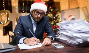 Mężczyzna w czapce Mikołaja pracuje w biurze do późna w Wigilię, otoczony stertami papierów i świątecznymi dekoracjami.