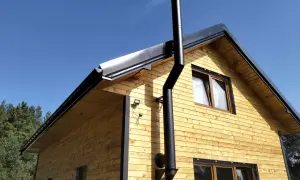Czarny, metalowy komin do kozy w domku letniskowym, zamontowany na drewnianej ścianie pod błękitnym niebem.