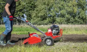 Mężczyzna w rękawiczkach i gumowcach obsługuje czerwoną glebogryzarkę na polu.