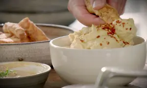 En hand doppar en bit i krämig hummus toppad med chiliflakes. Bredvid står en skål med gyllene halloumi fries.