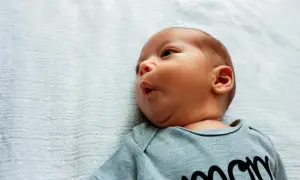 Uroczy niemowlak z kręczem szyi, ubrany w body z napisem "mom", leży na jasnoniebieskim tle.