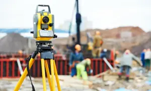Żółty reper geodezyjny na statywie na placu budowy. W tle pracownicy w kaskach i maszyny budowlane.