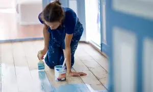 Mujer con mono azul se arrodilla para pintar el suelo de madera de azul claro. Se puede pintar el suelo para renovar la casa.
