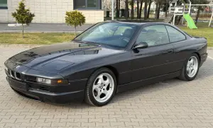 Czarne BMW V12, klasyk z lat 90., zaparkowane na kostce brukowej przed budynkiem z placem zabaw w tle.