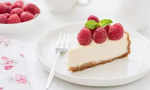 Una fetta di torta fredda allo yogurt guarnita con lamponi freschi e menta, su un piatto bianco con una forchetta.