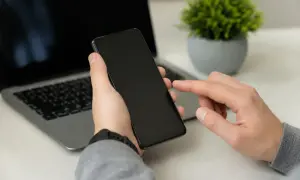 Dłoń trzyma smartfon, palec wskazujący dotyka ekranu, przygotowując się do całkowitego czyszczenia telefonu. Obok laptop i roślina.