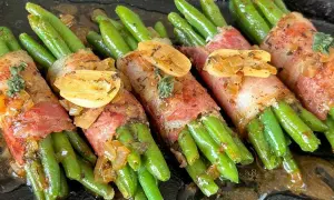 Grüne Bohnen im Speckmantel, mit Knoblauch und Kräutern verfeinert, liegen auf einer dunklen Platte in einer köstlichen Sauce.