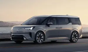Elegancki, szary minivan Volvo EM90 jedzie wzdłuż wybrzeża o zachodzie słońca.
