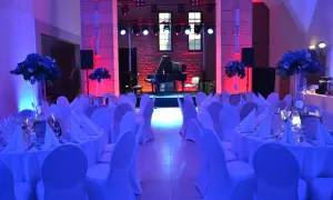 Elegancka filharmonia restauracja z fortepianem na scenie, stoły nakryte białymi obrusami i ozdobione kwiatami, oświetlone niebieskim i różowym światłem.