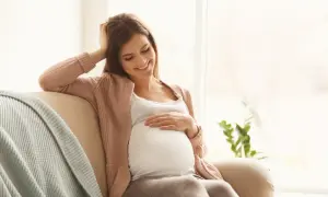 Uśmiechnięta ciężarna kobieta siedzi na kanapie, delikatnie dotykając brzucha. Myśli o bezpiecznym oczyszczaniu wodorowym w ciąży.