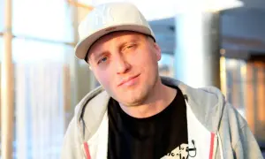 Młody raper w czapce z daszkiem i bluzie z kapturem, z lekkim uśmiechem na twarzy.