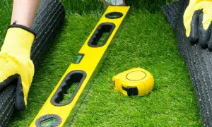Manos enguantadas preparan terreno para césped artificial. Nivel y cinta métrica listos para el proyecto.