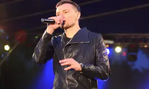 Artysta liber śpiewa do mikrofonu na scenie, ubrany w czarną kurtkę.