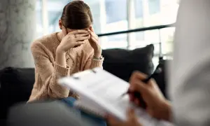 Kobieta w beżowym swetrze, z dłońmi na twarzy, przeżywa trudne emocje podczas terapii traumy. Terapeuta notuje.