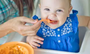 Ein Baby in einem blauen Kleid erhält Brei vom Löffel. Die Augen des Babys strahlen, ein Zeichen für die ersten reifezeichen baby.