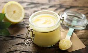 Un pot de lemon curd onctueux, une moitié de citron et des feuilles vertes sur fond de bois. Une cuillère en bois est prête à déguster.