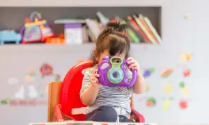 A young child in a striped shirt sits in a red high chair, holding a purple toy camera to their face.