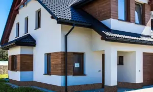 Nowy dom z białym tynkiem i drewnianymi akcentami. Brak cokołu na elewacji, widoczna kamyczkowa podmurówka.
