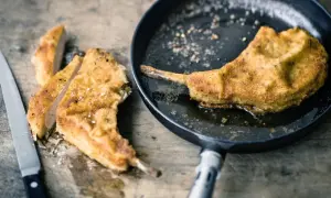 Ein paniertes Kotelett braten in der Pfanne. Daneben liegen bereits geschnittene Stücke auf einem Holzbrett, daneben ein Messer.