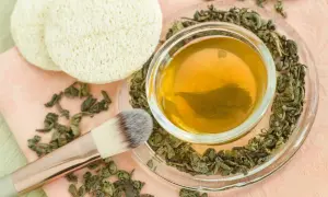 Naczynie z naparem zielonej herbaty, otoczone suszonymi liśćmi. Obok pędzel do makijażu i dwa okrągłe płatki. Zielona herbata na włosy - naturalny sekret piękna.