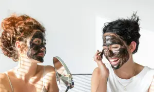 Dos mujeres con mascarilla negra en el rostro, una se mira en un espejo, buscando quitar espinillas negras grandes.