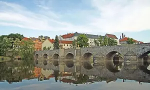 Historische Steinbrücke über den Fluss in Písek, Tschechien, mit Spiegelung im Wasser und bunten Häusern im Hintergrund.