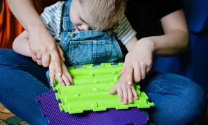 Dziecko w ogrodniczkach z pomocą terapeuty bawi się sensorycznymi matami. Terapia dla dzieci z problemami emocjonalnymi często wykorzystuje takie zabawy.