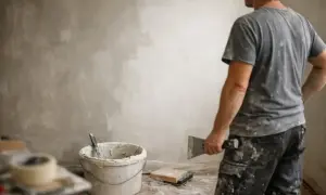 Pracownik z kielnią czeka, aż gładź na ścianie wyschnie. Obok wiadro z zaprawą i taśma malarska.